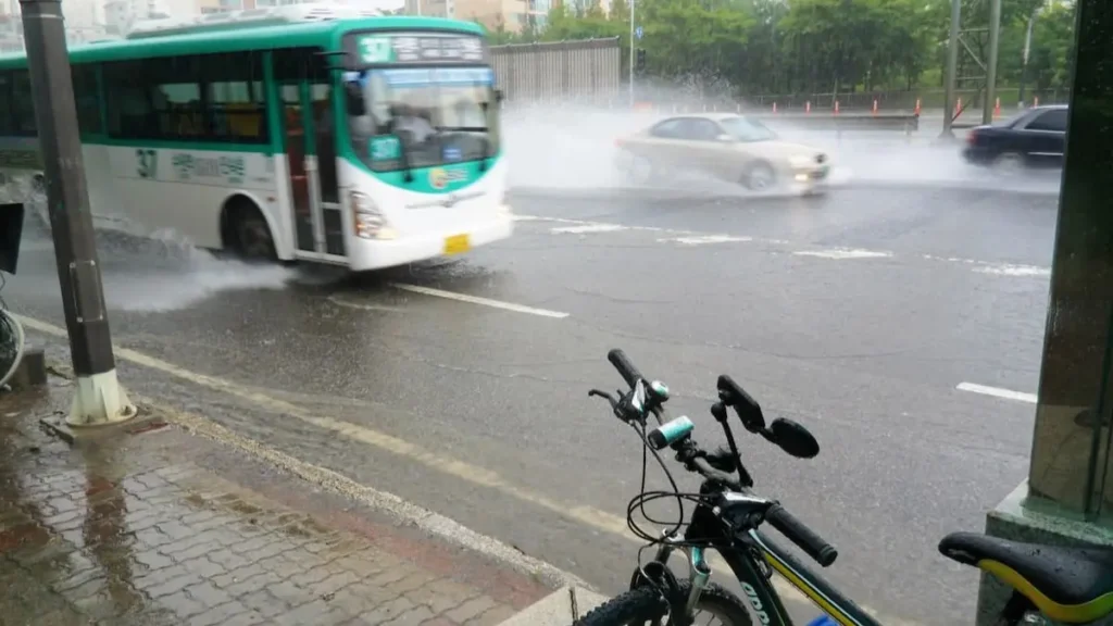폭우가 쏟아지는 도시의 도로, 버스가 물을 튀기며 지나가고 자전거가 서 있다.
