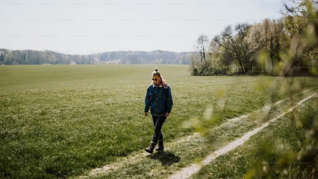 편하게 입은 여성이 이어폰이나 헤드폰 없이 햇살이 비추는 넓은 들판 길을 혼자 조용히 걷고 있는 모습