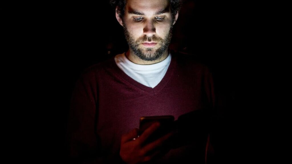 어두운 방 안에서 스마트폰 불빛에 비친 피곤한 얼굴, 밤새 미국 주식 차트를 보는 직장인의 모습