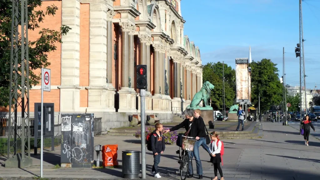 코펜하겐의 웅장한 박물관 건물 앞 인도에서 할머니와 손자, 손녀로 보이는 가족이 자전거 옆에 서서 다정하게 이야기를 나누는 모습. 주변의 사자 동상과 신호등이 보인다.